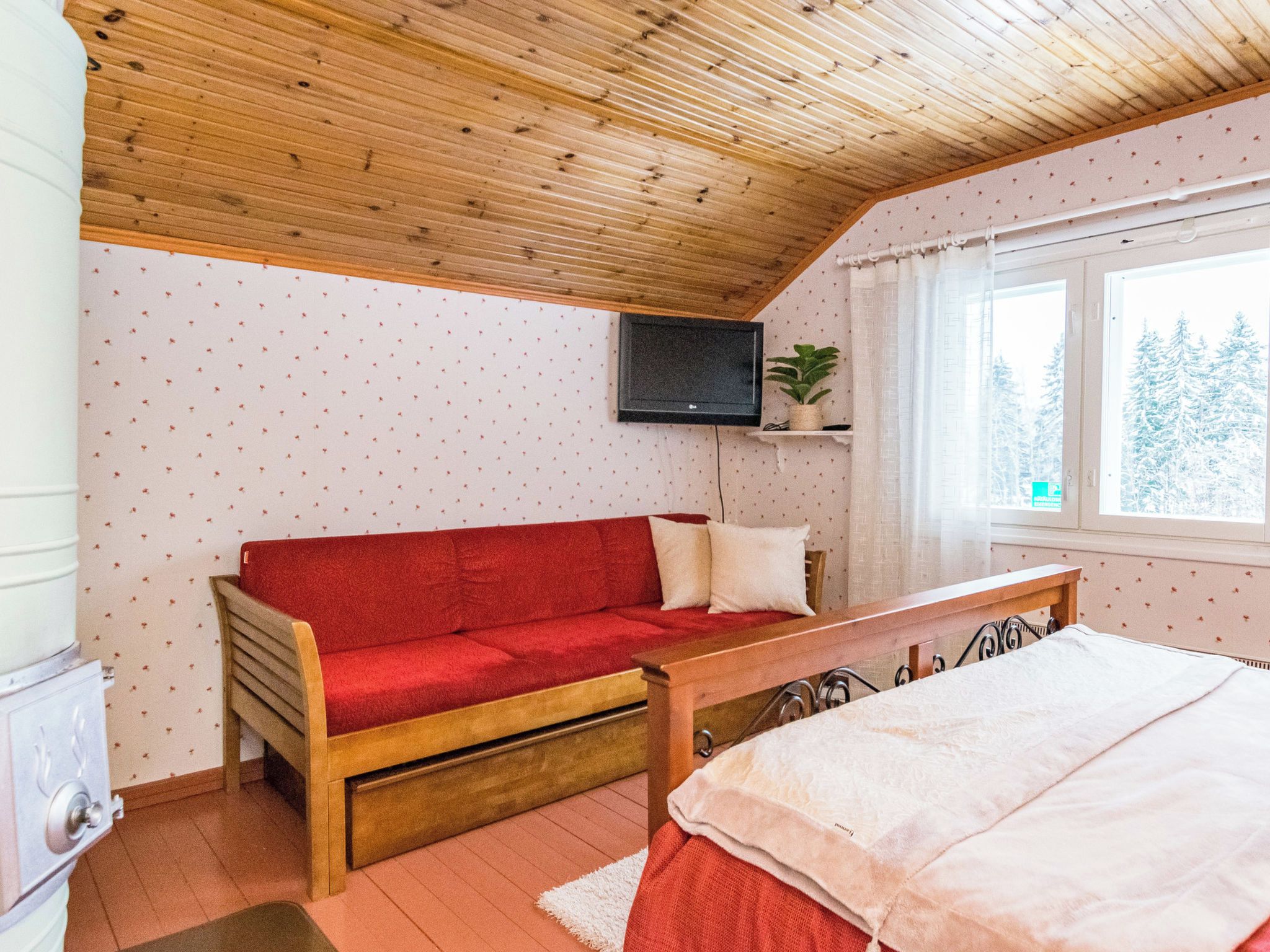 A cozy room with a wooden ceiling, a red sofa, a bed, a TV mounted on the wall, and a window with a view of trees.