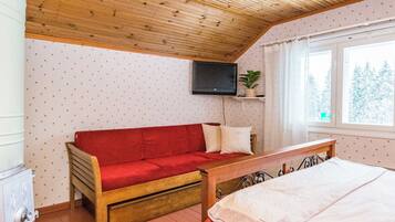 A cozy room with a wooden ceiling, a red sofa, a bed, a TV mounted on the wall, and a window with a view of trees.
