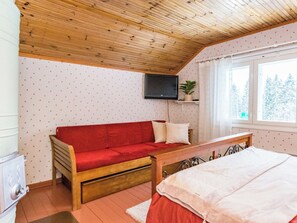 Een knus ingerichte kamer met een houten plafond, een rode bank, een bed, een aan de muur bevestigde televisie en een raam met uitzicht op bomen.
