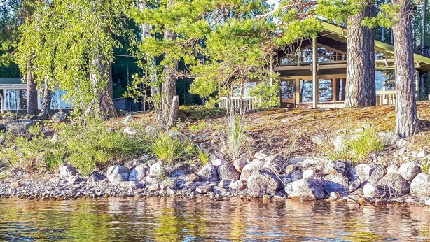Blick auf einen See mit einer Holzhütte, Bäumen und einer felsigen Uferlinie.