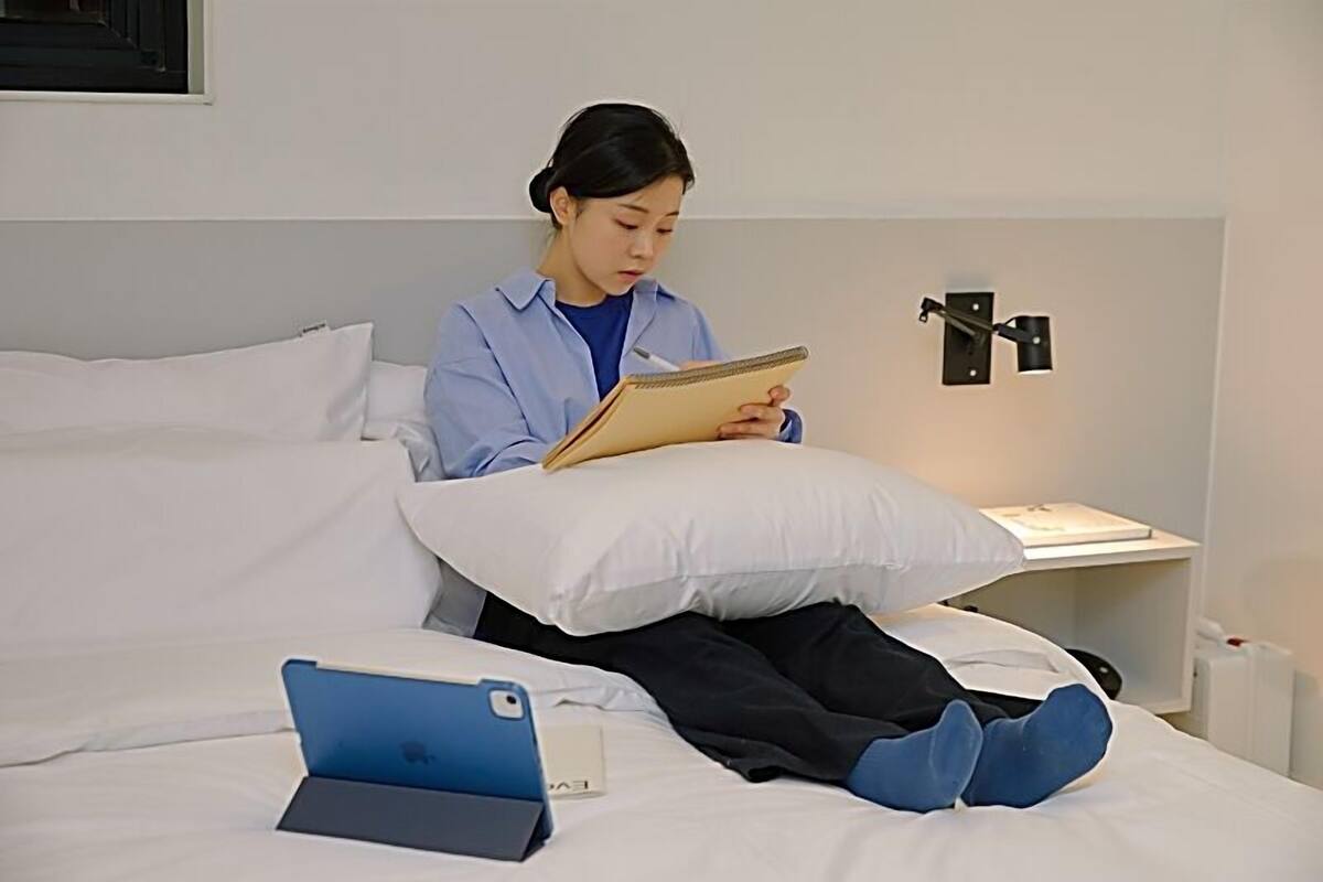Een persoon zittend op een bed met een notitieboek, een laptop en een kussen.