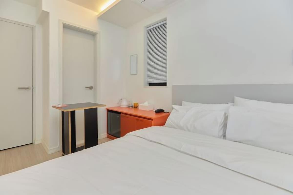 A bedroom with a bed, a nightstand, a small table, a window with blinds, and a door.