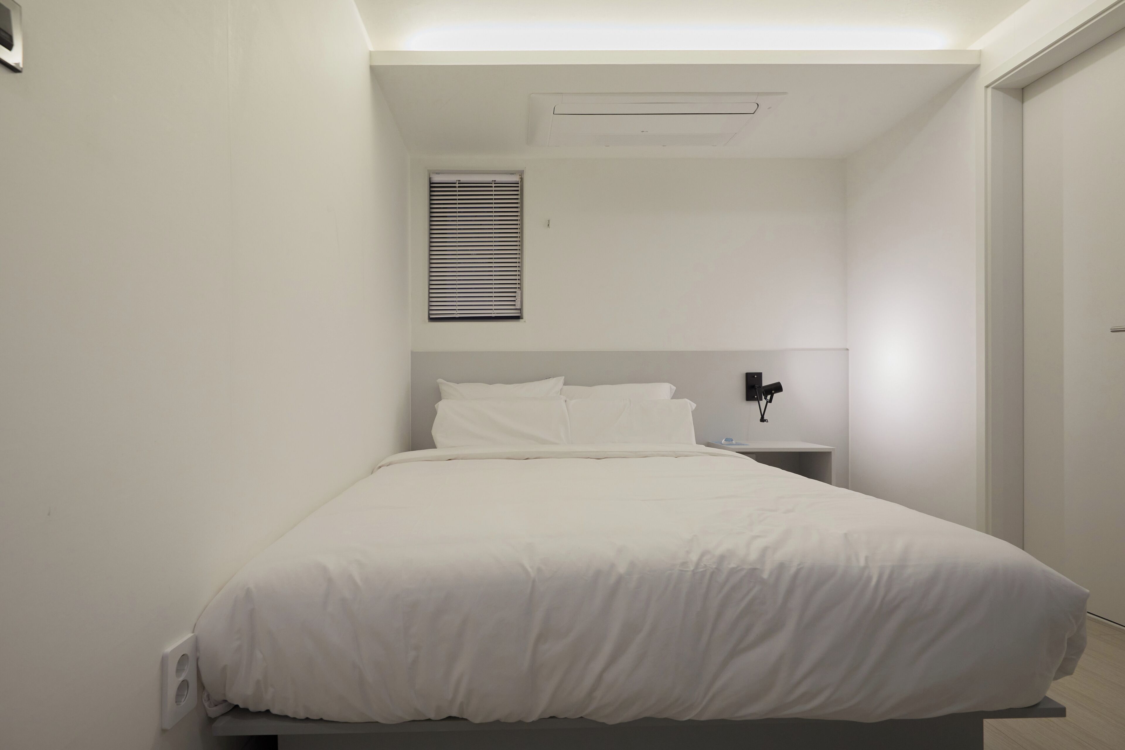 Une chambre à coucher minimaliste, avec un lit simple, une table de chevet, une lampe fixée au mur et une fenêtre avec des stores.