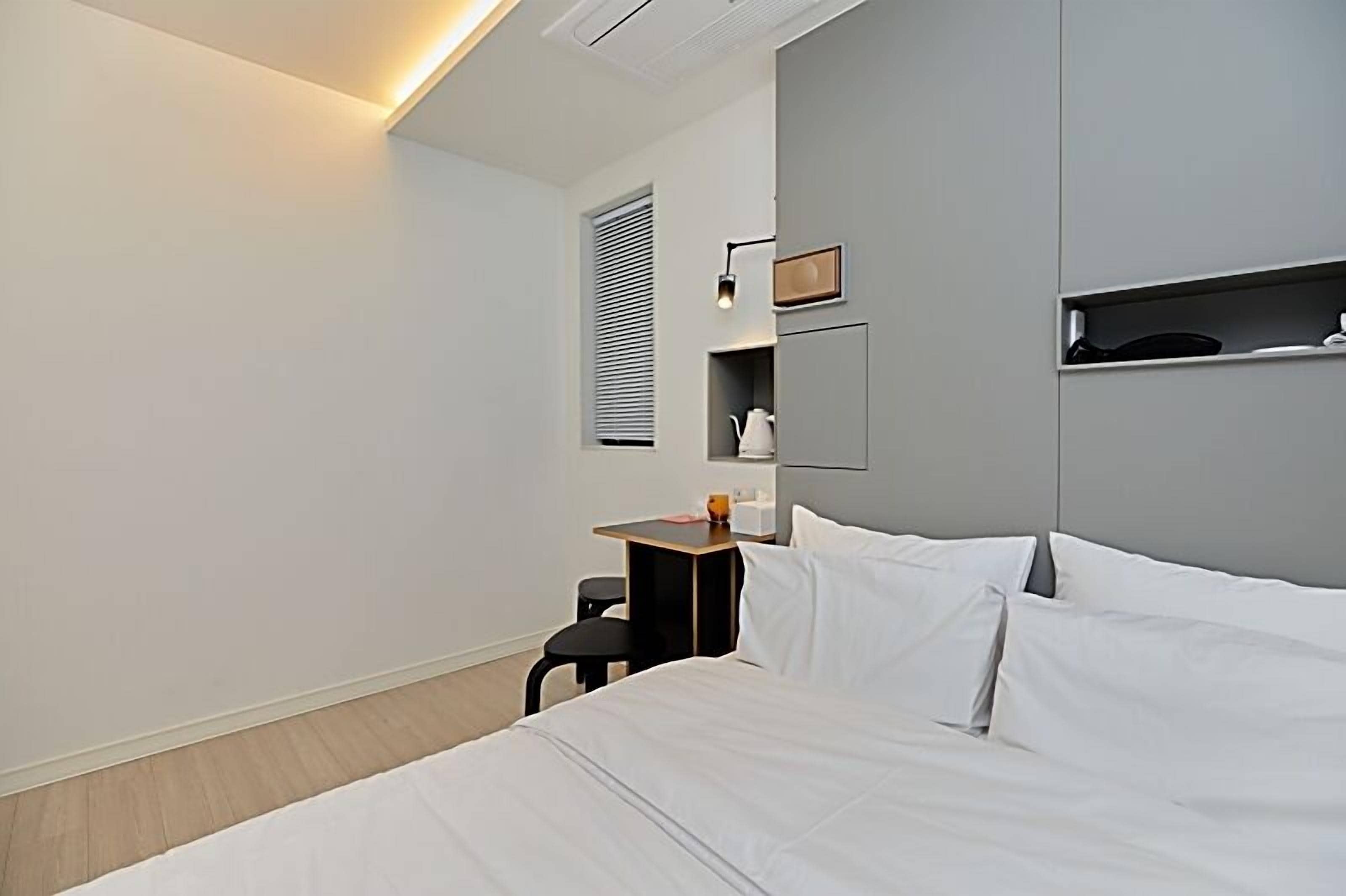 Et moderne hotelværelse med seng, lille bord, stol, natbord, vægmonteret hylde og et vindue med persienner.