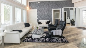 Een ruime woonkamer met een witte bank, een zwarte lederen fauteuil en een vloerkleed met dessin.