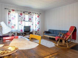 A living room with a wooden table, a rocking chair, a blue sofa, a television, and a window with floral curtains.