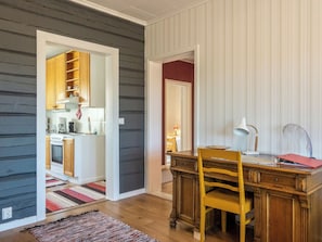 A room with a wooden desk, chair, and a kitchen area with cabinets and a stove.