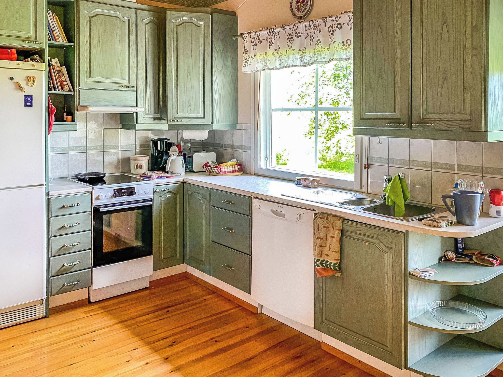 Een keuken met een fornuis, oven en vaatwasser, houten kastjes en een raam met uitzicht op groen.