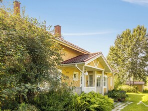 Une maison jaune avec un toit rouge, entourée d’une végétation luxuriante et sous un ciel bleu clair.
