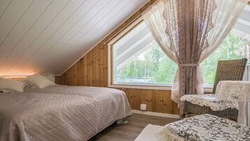 A bedroom with a sloped ceiling, a large window with curtains, a bed with a quilted cover, and a wicker chair with a table.