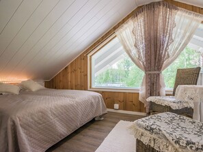 A bedroom with a sloped ceiling, a large window with curtains, a bed with a quilted cover, and a wicker chair with a table. - Kaislaranta by Interhome (Suonenjoki)