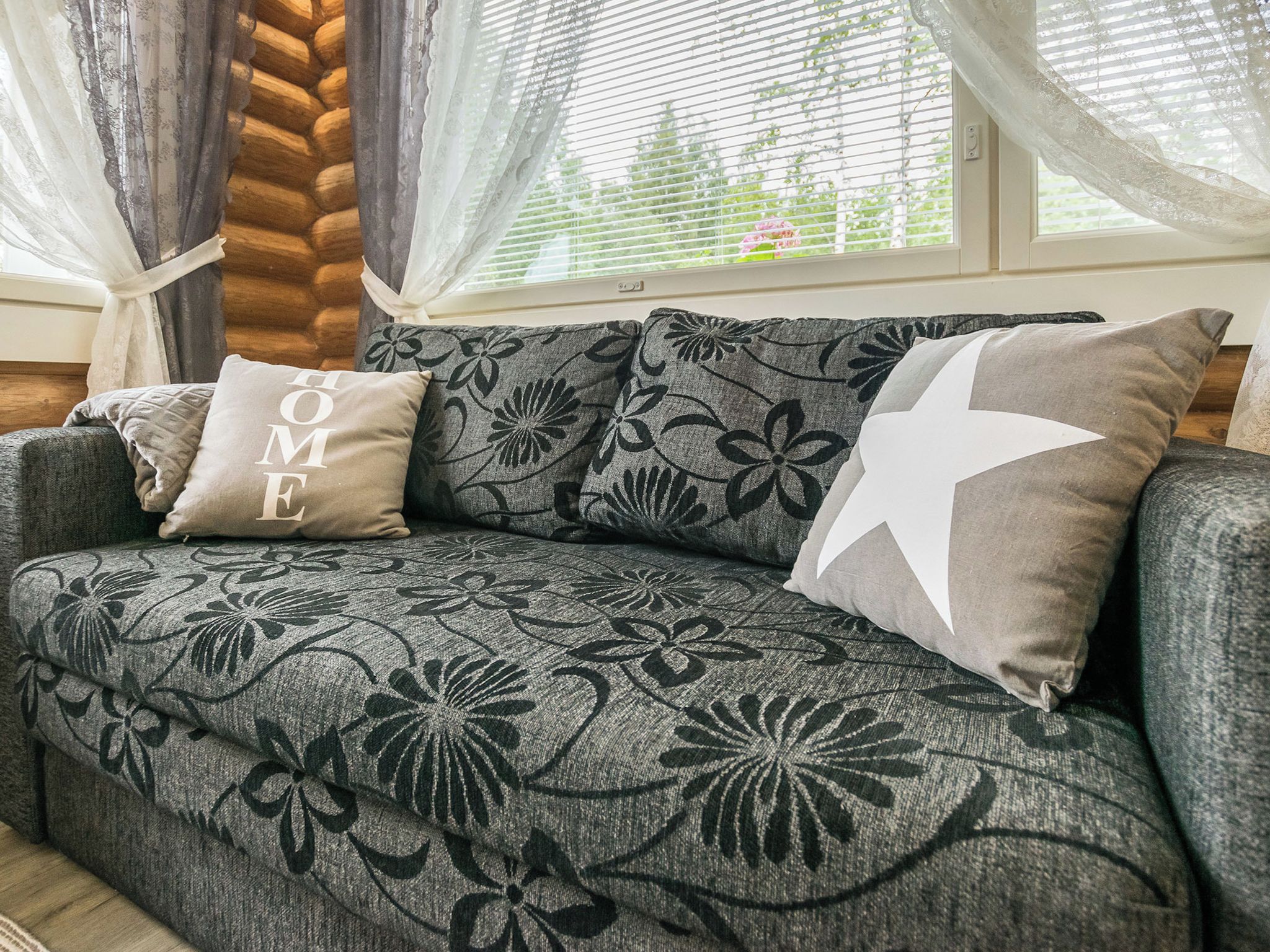 Un sofá gris con tapicería floral y dos almohadones, uno con el texto 'HOME', en una habitación con ventana y cortinas.