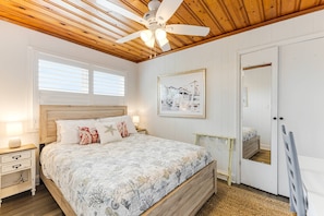 Un dormitorio con techo de madera, una cama con colcha estampada, una mesita de noche con lámpara, un cuadro enmarcado en la pared y una puerta con espejo.
