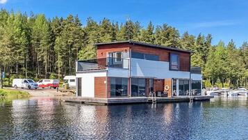 Une maison moderne à deux étages, dotée d’un quai, entourée d’une forêt et bordée par un plan d’eau.