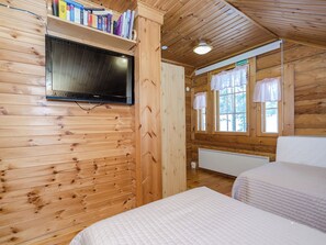 A wooden cabin room with a television mounted on the wall, a bed, a window with curtains, and a shelf with books. - Villa tunturitervakko by Interhome (Liperi)