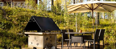 A rustic outdoor grill setup with a stone structure, surrounded by a dining table and chairs, in a forested area with a lodge in the background.