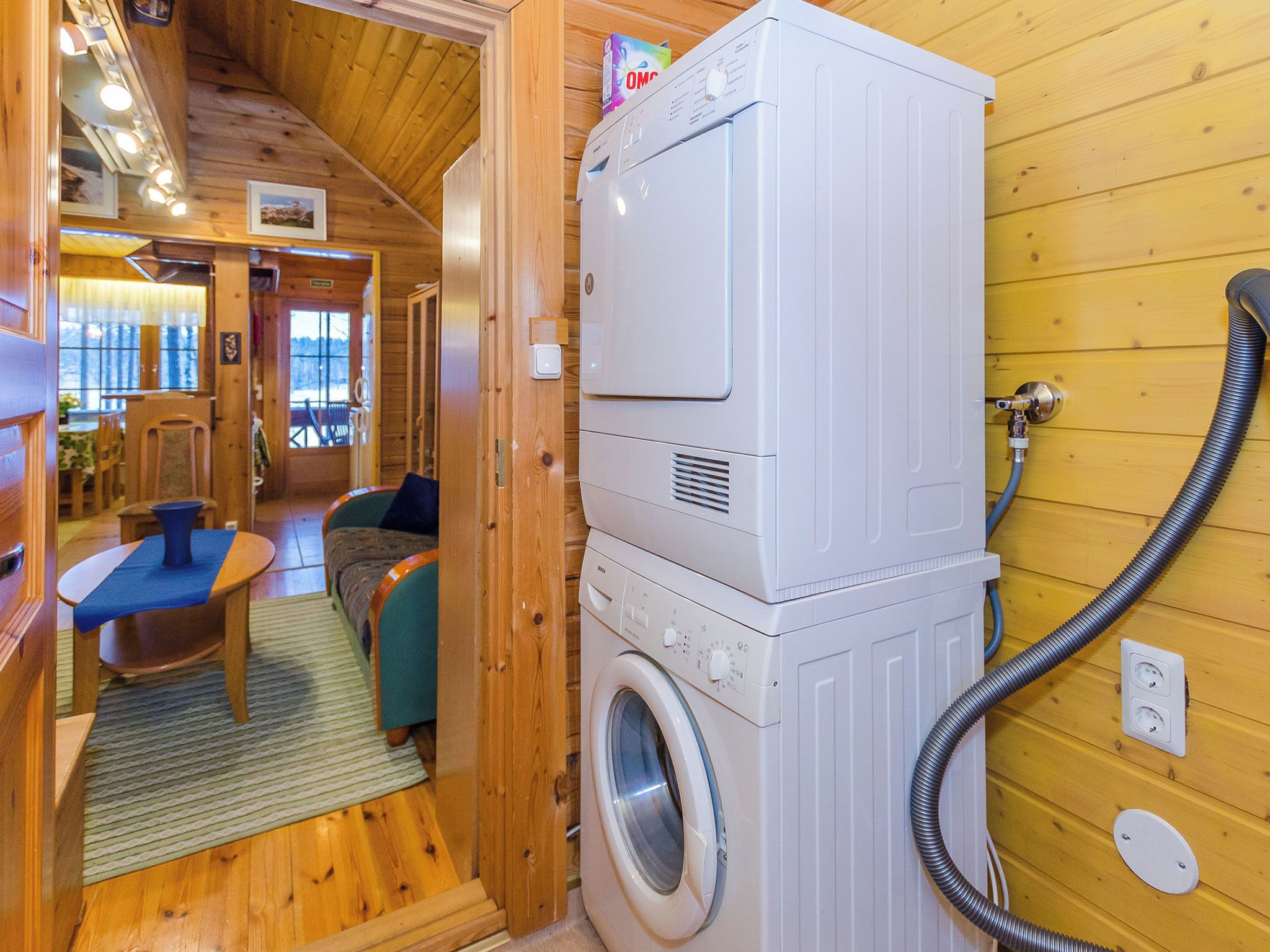 A compact room with a washing machine and a hose, wooden walls, a small seating area with a table and chairs, and a window with a view.