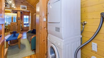 A compact room with a washing machine and a hose, wooden walls, a small seating area with a table and chairs, and a window with a view.