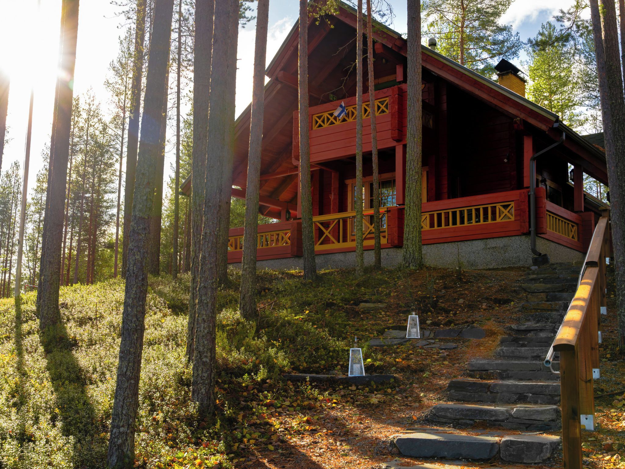 Une cabane en bois à l’extérieur rouge et aux accents jaunes, entourée d’une forêt, avec un escalier en pierre menant à l’entrée.