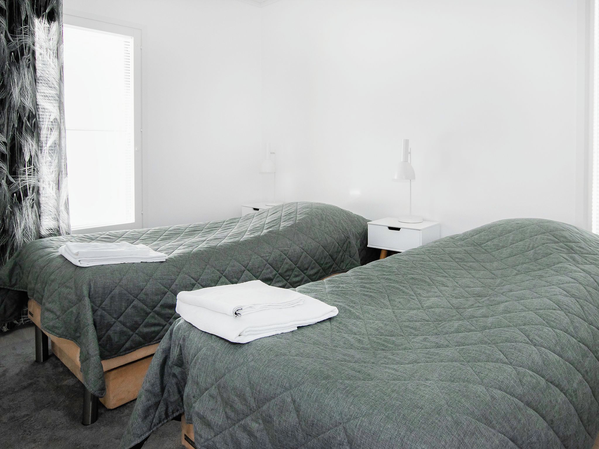 A twin room with two beds, each with a green quilted bedspread and white towels. There are bedside tables with lamps and a window with curtains.
