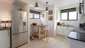 Une cuisine moderne avec un réfrigérateur en acier inoxydable, une table en bois avec des tabourets et une fenêtre dotée d’un store vert.