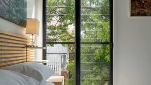 Une chambre à coucher avec un lit, une table de chevet, une lampe et une grande fenêtre donnant sur les arbres.