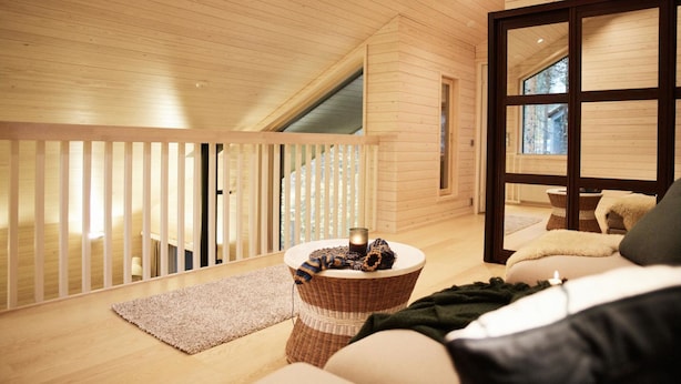 Un espacioso dormitorio tipo loft con techo alto, vigas de madera, un ventanal grande y un rincón de descanso acogedor con una mesita de mimbre.