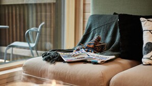 Un salon chaleureux avec un canapé, un magazine, une bougie et une fenêtre donnant sur une chaise à l’extérieur.