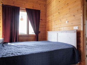 A room with wooden walls, a bed with a quilted cover, a radiator, and curtains on a window.