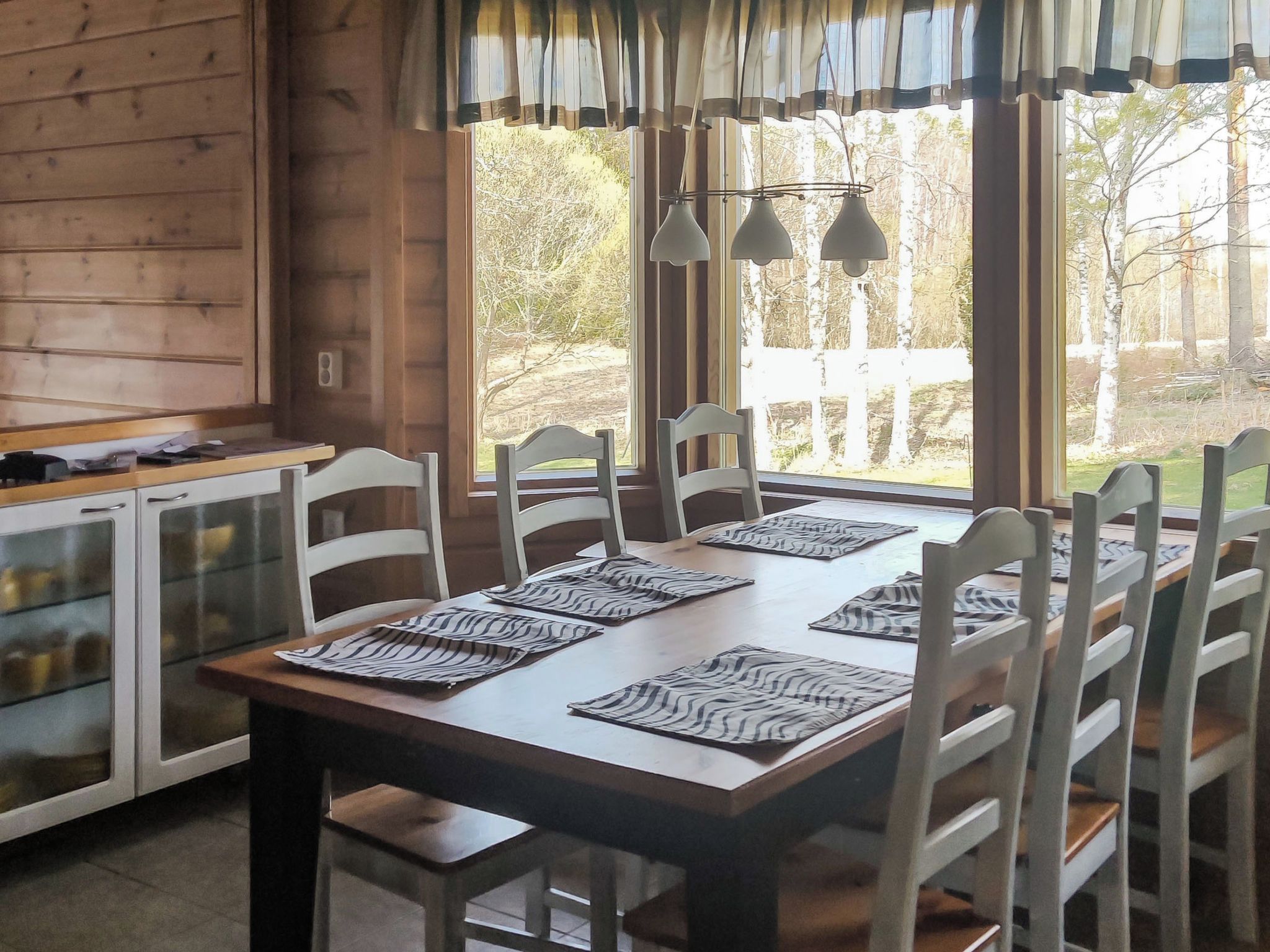Un intérieur en bois comprenant une table à manger pour six personnes, des chaises, un coin cuisine et une vue sur les arbres à travers de grandes fenêtres.