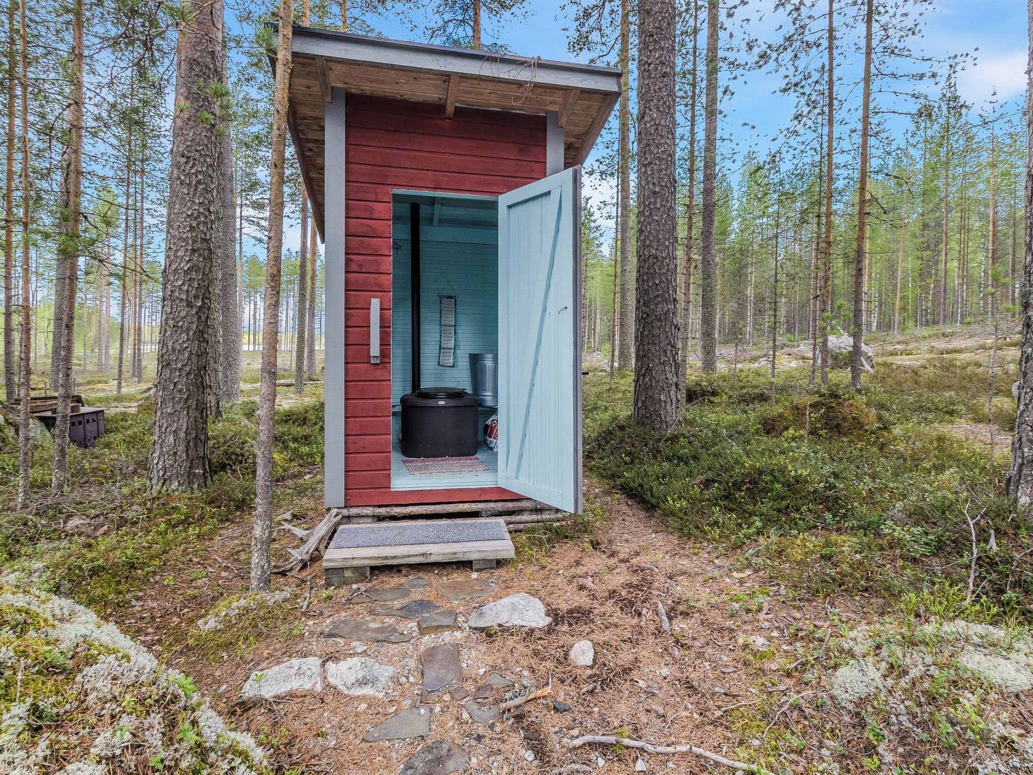 A small cabin with a red exterior and a blue door, surrounded by a forest.
