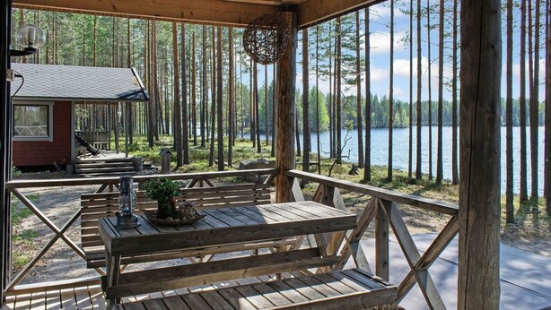 Une terrasse en bois avec des tables de pique-nique et des bancs, donnant sur un lac et entourée de grands arbres.