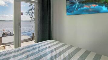 A bedroom with a bed, a window with a view of a lake and mountains, and a painting of a mountain landscape.