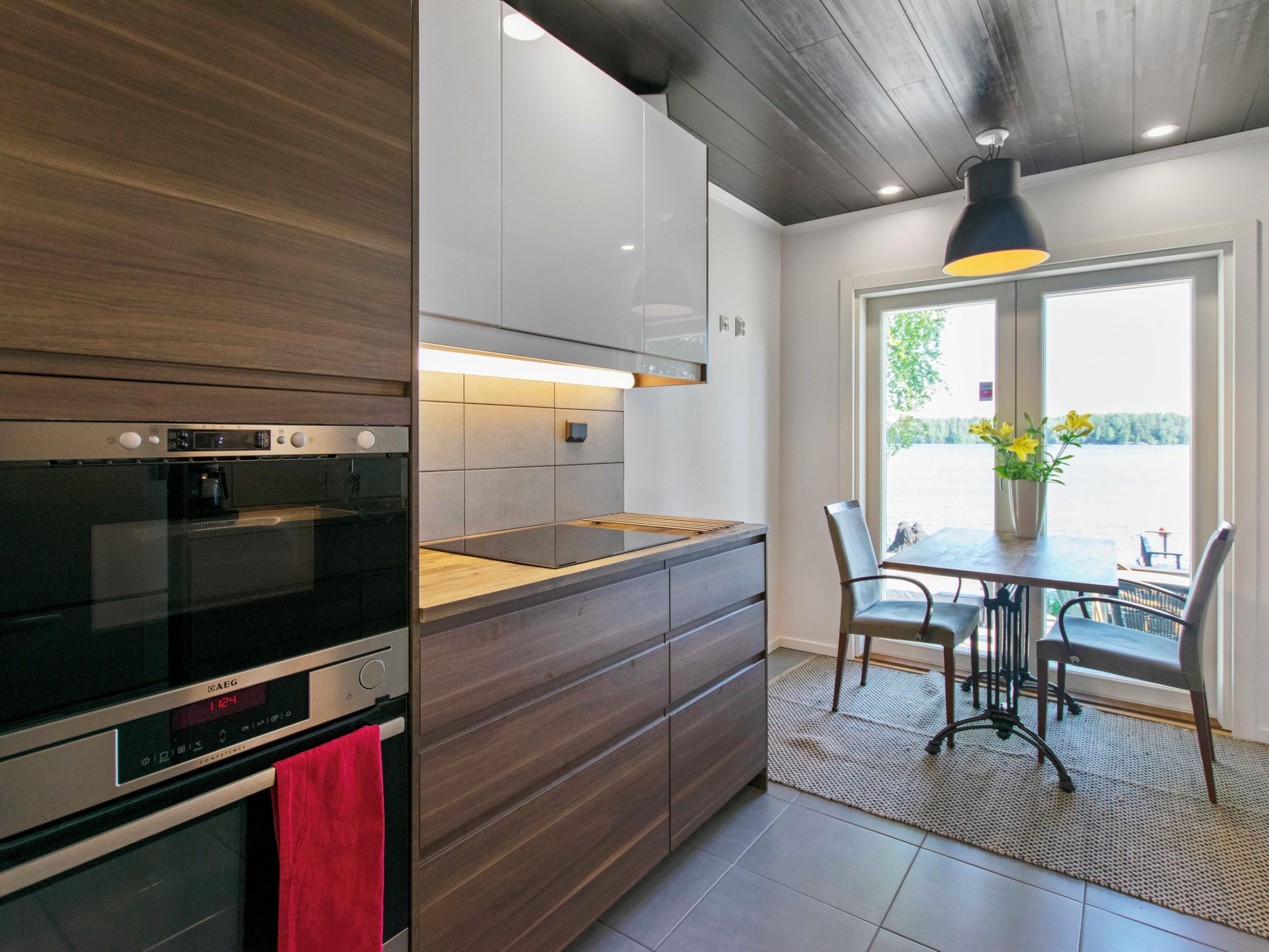 Une cuisine moderne dotée d’un four encastré, un coin repas avec une table et des chaises, et une vue sur un plan d’eau par le biais d’une grande fenêtre.