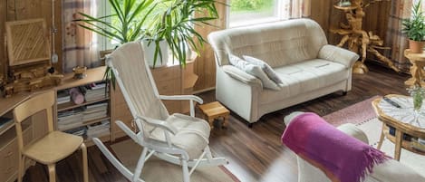 A cozy living room with a rocking chair, a sofa, and a coffee table. There is a large plant and a chandelier.