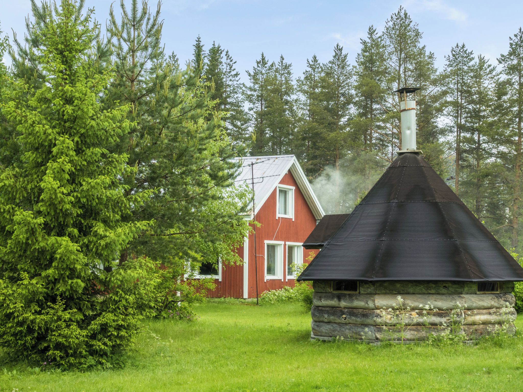 A rustic cabin with a chimney emitting smoke, surrounded by dense forest and a red house in the background.