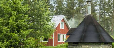 Een rustieke chalet met schoorsteen waar rook uit komt, omgeven door dicht bos en een rood huis op de achtergrond.