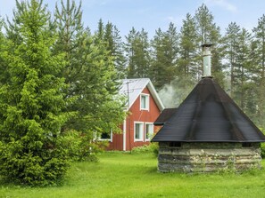 A rustic cabin with a chimney emitting smoke, surrounded by dense forest and a red house in the background.