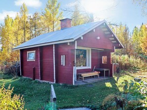 A small, red wooden cabin with a porch and a chimney, surrounded by trees and grass.