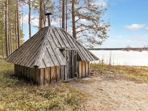 Una cabaña de madera rústica con techo inclinado y chimenea, ubicada en zona boscosa con un lago helado al fondo.