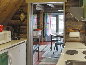 Een landelijke keuken met een houten plafond, een klein zithoekje met een tafel en stoelen, en een uitzicht door een deur naar een slaapkamer met een bed en gordijnen.