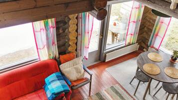 A cozy interior with a red sofa, a small table with woven placemats, and a view through large windows with colorful curtains.