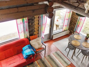 Ein gemütliches Interieur mit einem roten Sofa, einem kleinen Tisch mit gewobenen Untersetzern und Blick durch große Fenster mit bunten Vorhängen.