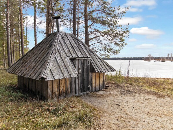 A rustic wooden cabin with a sloped roof, situated in a forested area with a frozen lake in the background.
