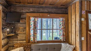 Ein gemütliches Blockhauszimmer mit Holzvertäfelung, einer Couch mit gemusterten Kissen, einem an der Wand montierten Fernseher und einem Fenster mit Vorhängen.