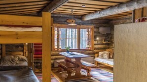 Ein rustikales Blockhaus-Interieur mit Holzbalkendecke, einem Esstisch mit Bänken, einer Couch und einem Fenster mit Vorhängen.