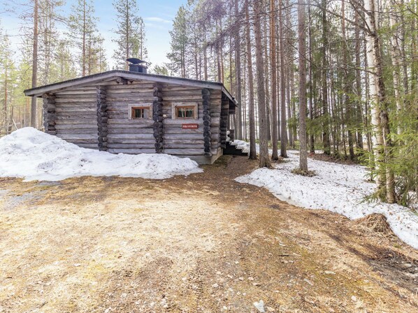 Une cabane en rondins avec une cheminée, une fenêtre et une porte, entourée d’une forêt et de neige.