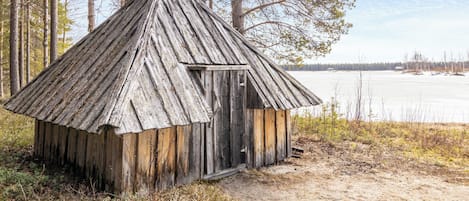 A rustic wooden cabin with a sloped roof, surrounded by trees and a frozen lake in the background.