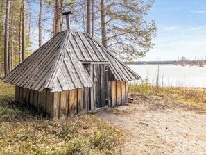 A rustic wooden cabin with a sloped roof, surrounded by trees and a frozen lake in the background.