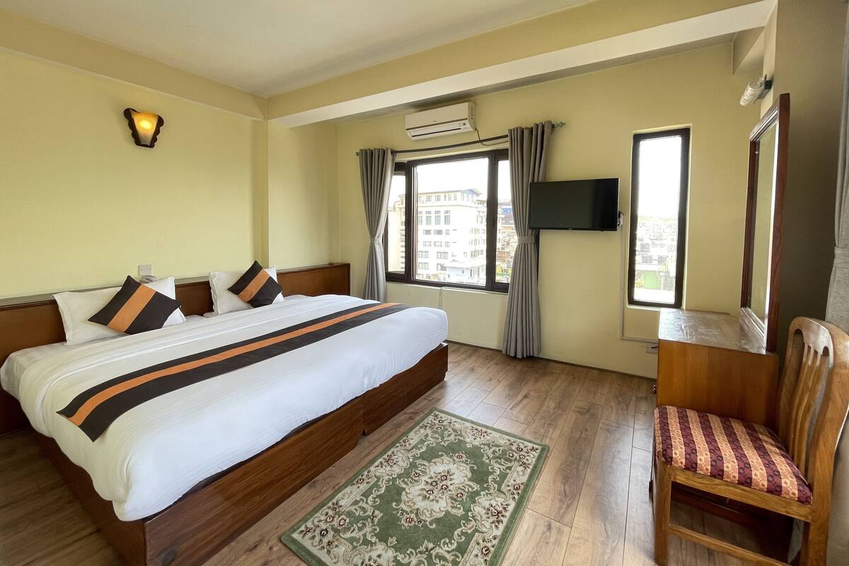 Una habitación de hotel con una cama grande, una silla de madera, un televisor montado en la pared y una ventana con cortinas.